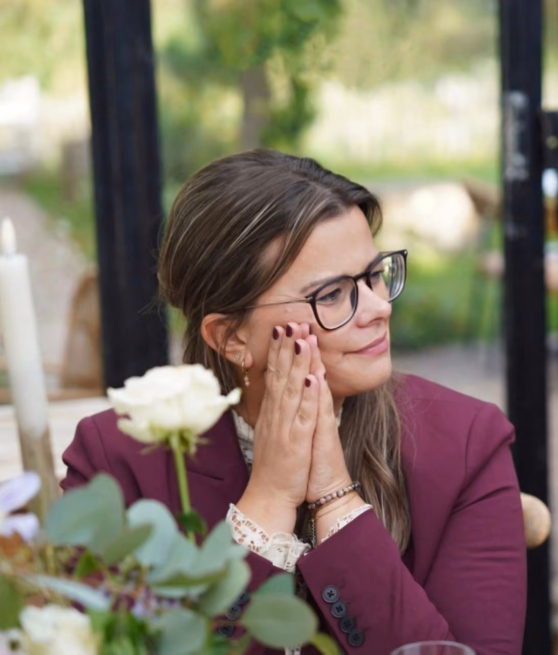 Nathalie zit aan een tafel met haar handen tegen elkaar bij haar wang en haar ogen zacht gericht. Op de voorgrond een witte roos en een kaars.