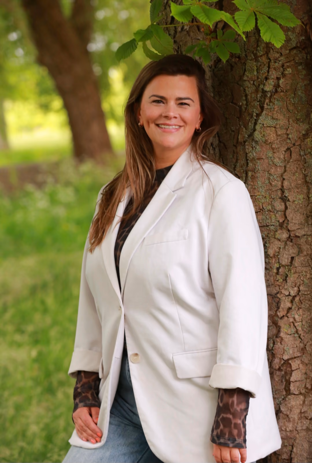Nathalie lacht in de camera. Ze staat in een lichte blazer tegen de stam van een kastanjeboom, met gras en bomen achter haar.
