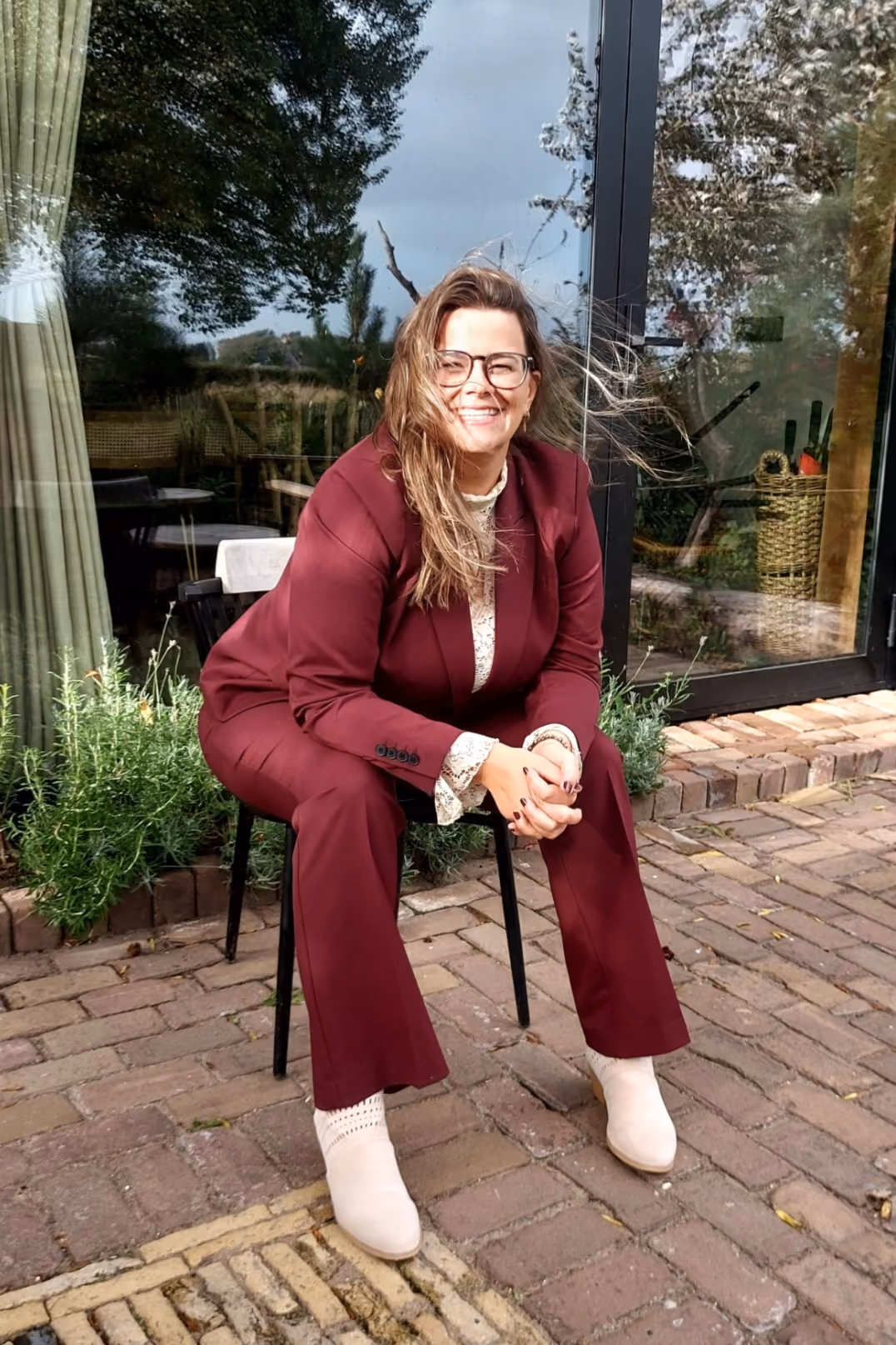 Nathalie zit lachend op een kruk voor een grote glazen pui, met haar haren in de wind. Naast haar een border met lavendel, onder haar een vloer van kleine klinkers.